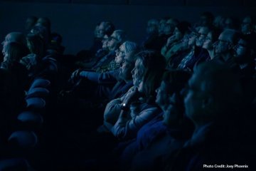 A side view of a group of people sit in a theater staring at a screen