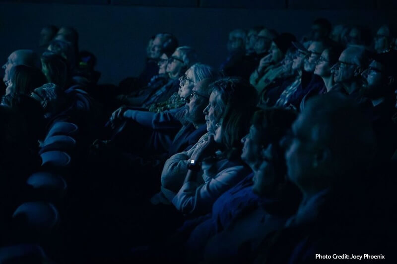 A side view of a group of people sit in a theater staring at a screen