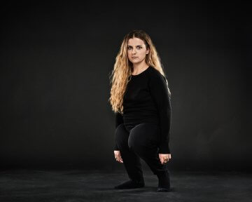 A blonde woman of short stature wearing black clothes stands against a black backdrop