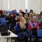 A group of hijabi women sit on blue chairs, observing something happening in front of them, some of them laughing, some keeping a straight face